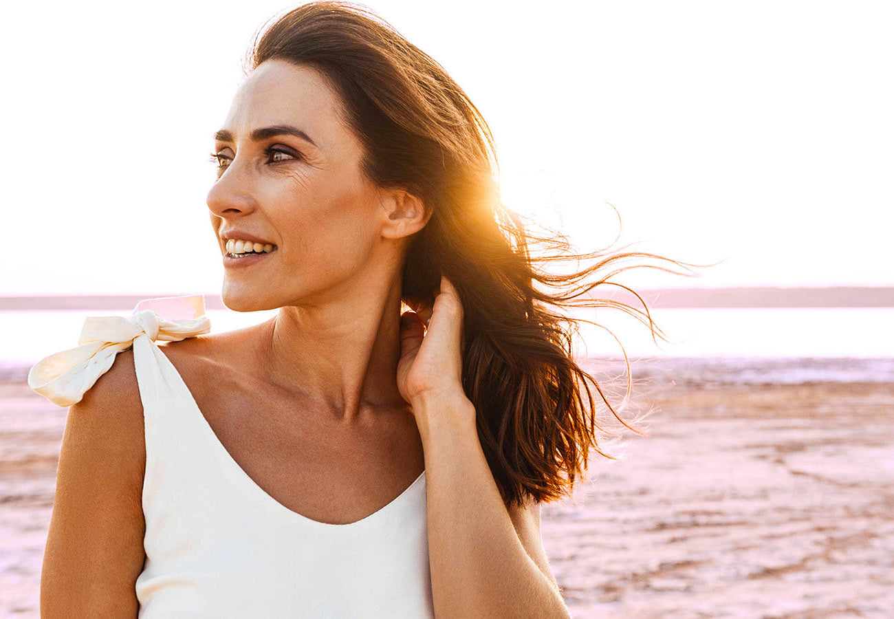 Jolie femme brune avec une belle peau repulpée et de beaux cheveux, devant un coucher de soleil, illustrant les bienfaits du Booster Collagène des Boosters Naturels pour la peau et la beauté des cheveux