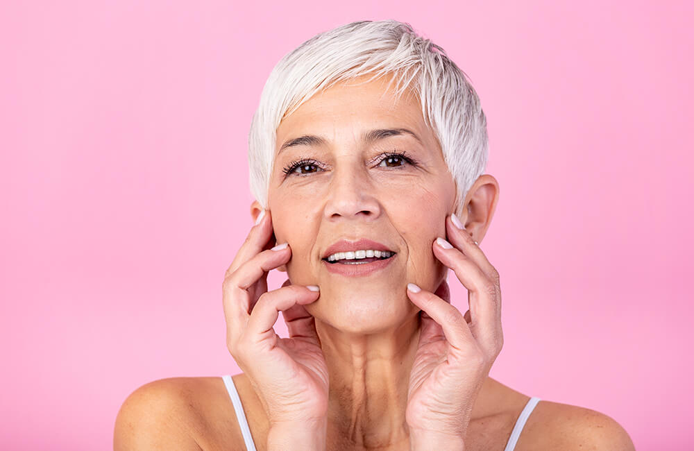 Femme mature souriante avec une belle peau, tenant son visage dans ses mains, illustrant les bienfaits du Booster de Collagène des Boosters Naturels pour hydrater la peau et favoriser le bien-être à tout âge