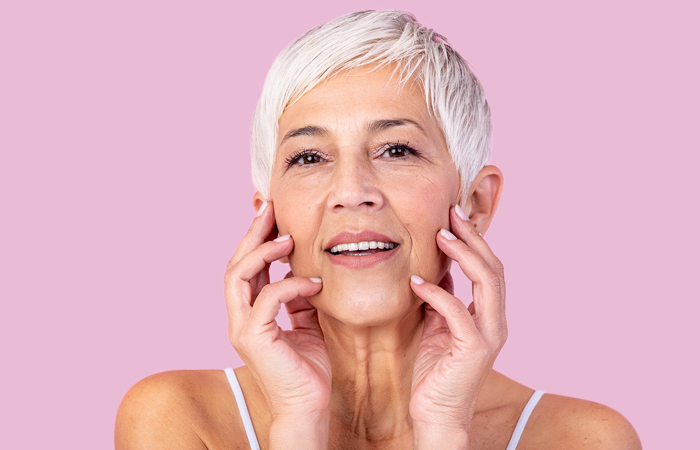 Femme mature avec des cheveux blancs tenant son visage, illustrant les bienfaits du Booster Collagène des Boosters Naturels pour une peau repulpée, hydratée, et anti-âge sur fond rose