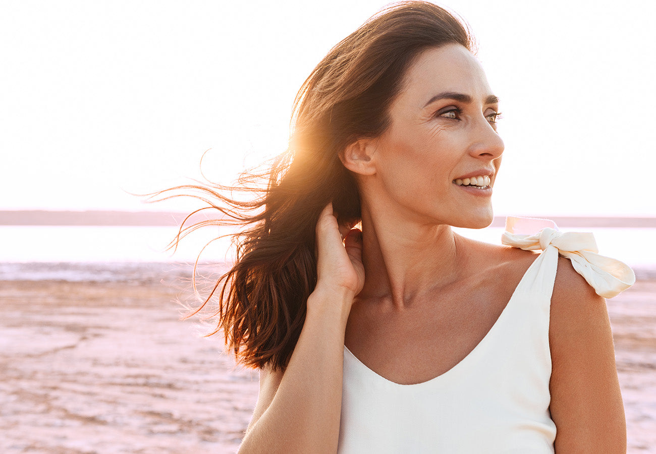 Femme de profil avec une belle peau et de beaux cheveux souriant devant un coucher de soleil, représentant le bien-être et les effets naturels des compléments alimentaires Les Boosters Naturels pour la peau et la beauté